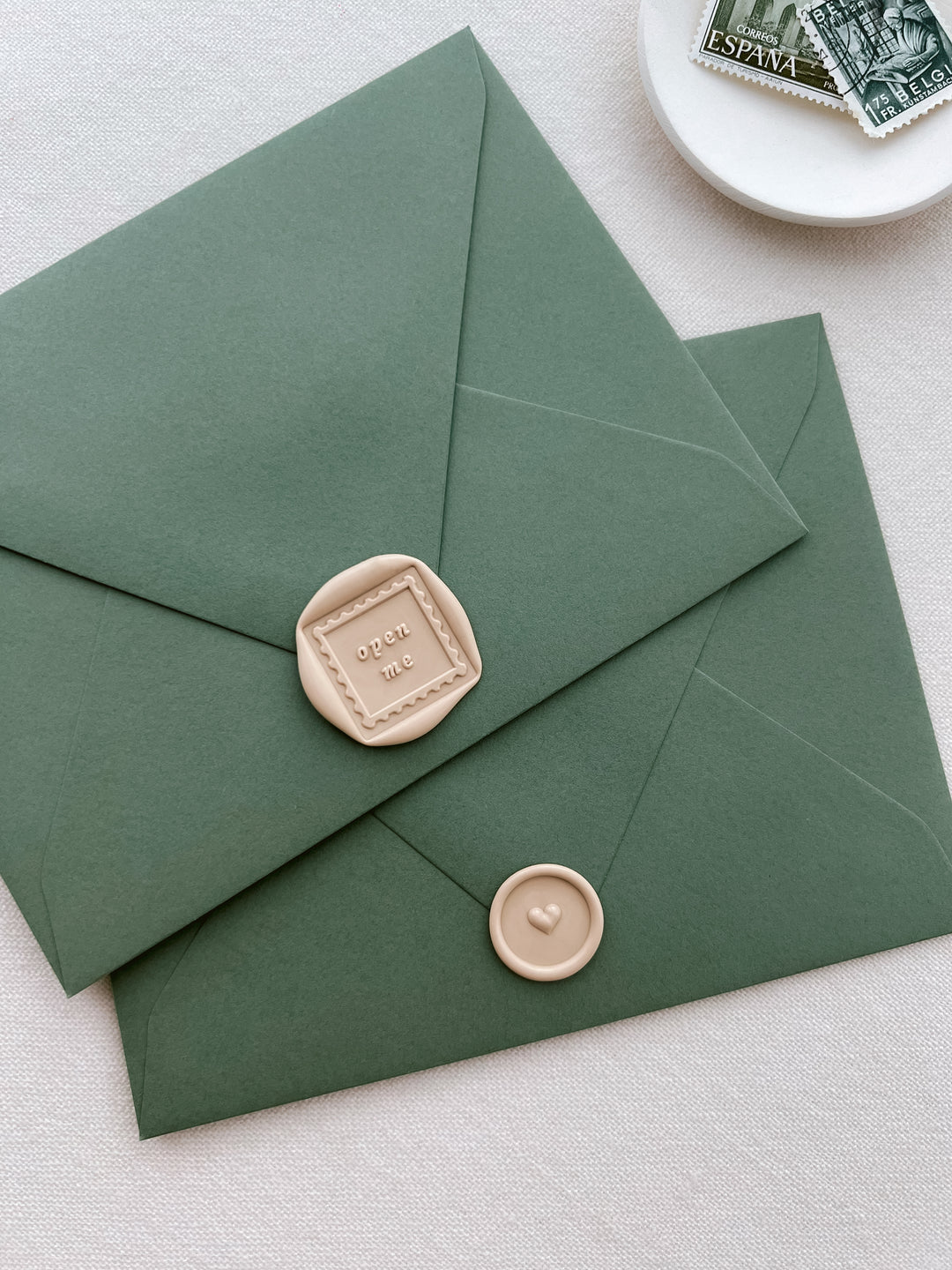 two beige wax seals on olive green envelopes, one featuring a postage stamp design with the phrase open me, and the other a 3D heart design mini wax seal 