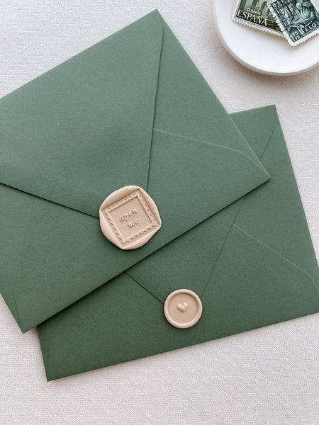 two beige wax seals on olive green envelopes, one featuring a postage stamp design with the phrase open me, and the other a 3D heart design mini wax seal 