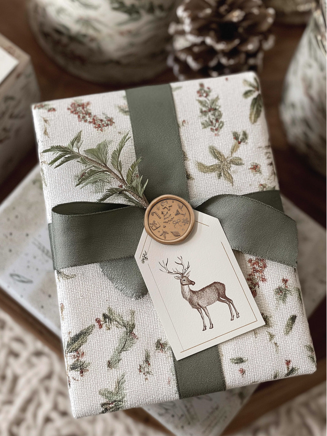 Gold Christmas wax seal featuring festive symbols such as snowflakes, candy cane, gingerbread man, Christmas tree, and more, on a wrapped holiday gift decorated with green silk ribbon and reindeer gift tag