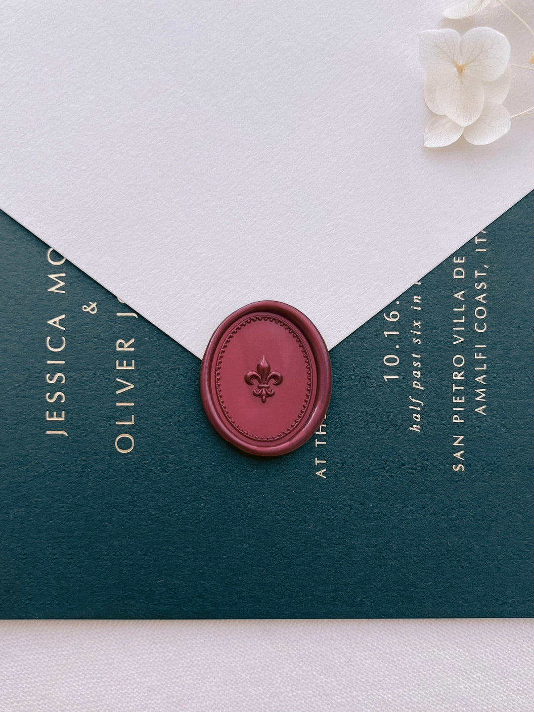 Dark metallic red 3D fleur-de-lis oval wax seal on a white envelope over a dark green gold foil wedding invitation card 