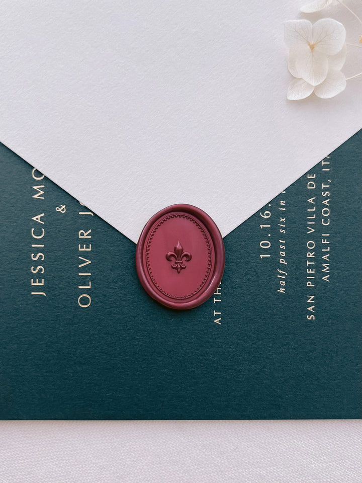 Dark metallic red 3D fleur-de-lis oval wax seal on a white envelope over a dark green gold foil wedding invitation card 