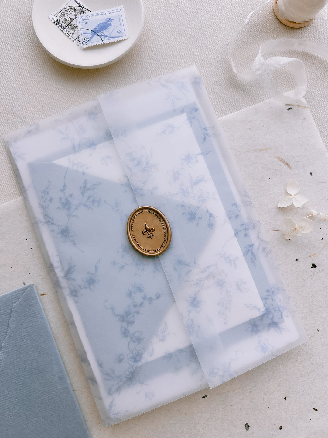 Bronze 3D fleur-de-lis oval wax seal on a blue botanical print vellum jacket over a wedding invitation suite 