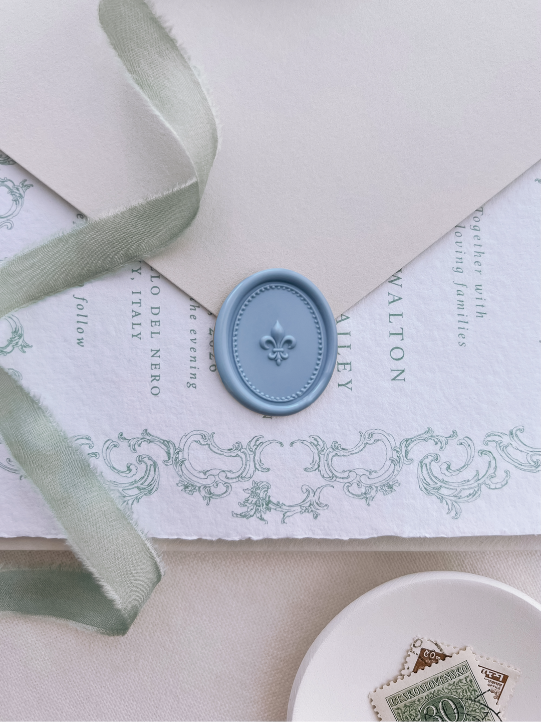 Blue oval 3D fleur-de-lis wax seal on a light beige envelope flap over a vintage-inspired white handmade paper wedding invitation card