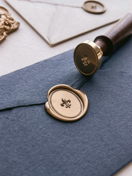 Black envelope with a gold oval wax seal featuring a fleur-de-lis design with a border design