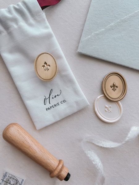 Fleur-de-lis brass stamp head with a wooden stamp handle, next to off-white and gold Fleur-de-lis oval wax seals