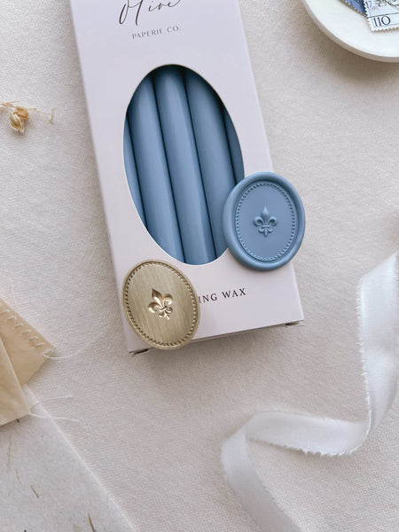 Dusty blue sealing wax sticks in a beige box with a dusty blue oval Fleur-de-lis wax seal placed on top