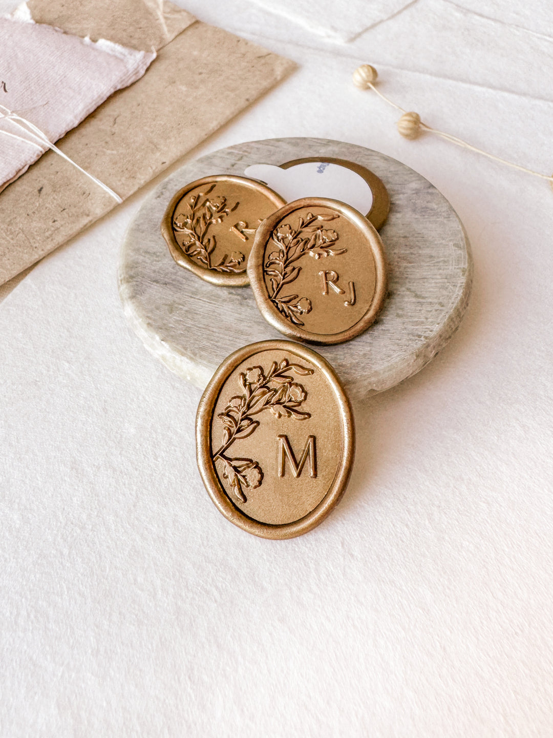 Oval floral monogram custom wax seals in gold styled with a small gray stone dish, handmade paper and a dried floral branch