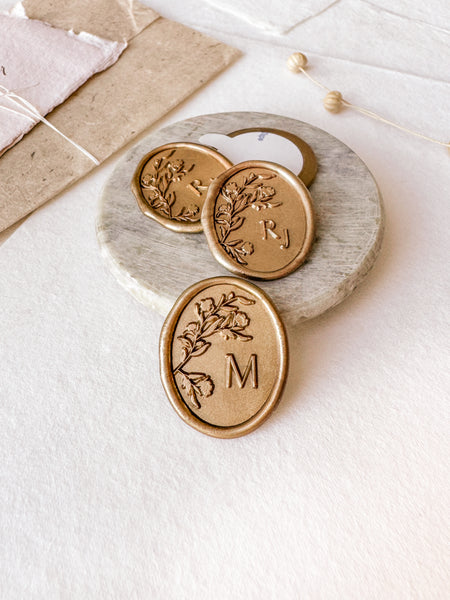 Oval floral monogram custom wax seals in gold styled with a small gray stone dish, handmade paper and a dried floral branch