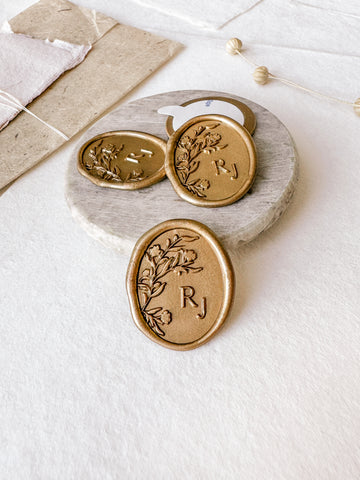 Oval floral monogram custom wax seals in gold styled with a small gray stone dish, handmade paper and a dried floral branch