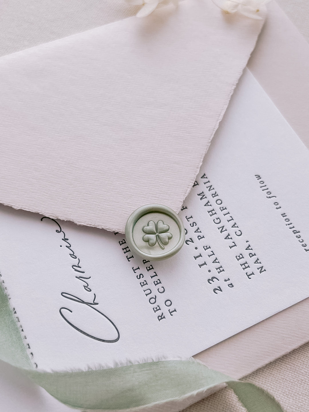 Light metallic sage green mini 3D four leaf clover wax seal on a light pink envelope over a wedding invitation card