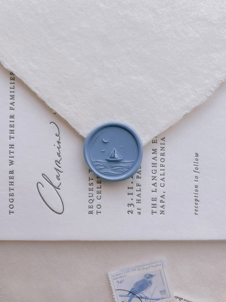 Blue nautical themed wax seal featuring a design of a sailboat over ocean waves under a crescent moon on white wedding invitation card and envelope