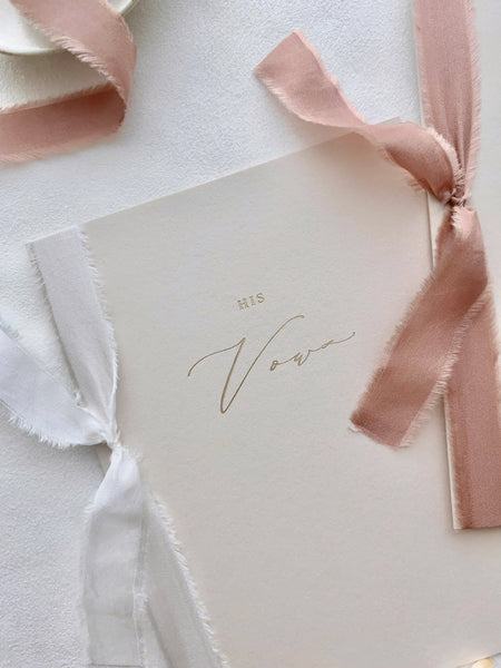 A set of two His and Her Vows beige card stock vow books in calligraphy script printed in gold foil one tied in dusty rose colored silk ribbon and one tied in white colored silk ribbon