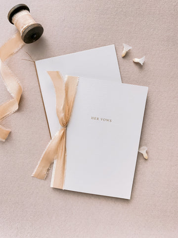 A set of two His and Her gold foil beige card stock vow books in typeface font with one tied in light gold colored silk ribbon and the other in fine brown twine