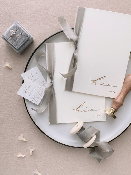 A set of two His and Her gold foil ivory card stock vow books in calligraphy script with taupe silk ribbon styled with bride wedding rings, spool of taupe silk ribbon and Olive Paperie Co postage style stamps