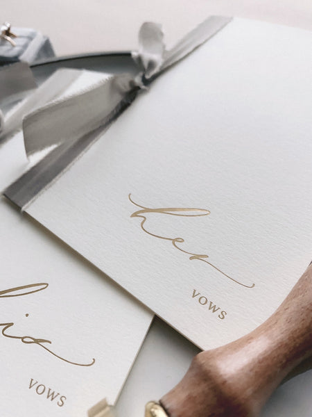 A set of two His and Her gold foil ivory card stock vow books in calligraphy script with taupe colored silk ribbon