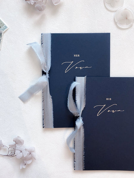 A set of two His and Her gold foil navy card stock vow books in calligraphy script with pale blue colored silk ribbon