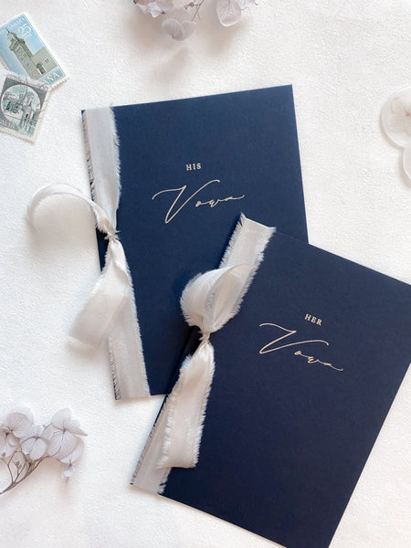 A set of two His and Her gold foil navy card stock vow books in calligraphy script with nude colored silk ribbon