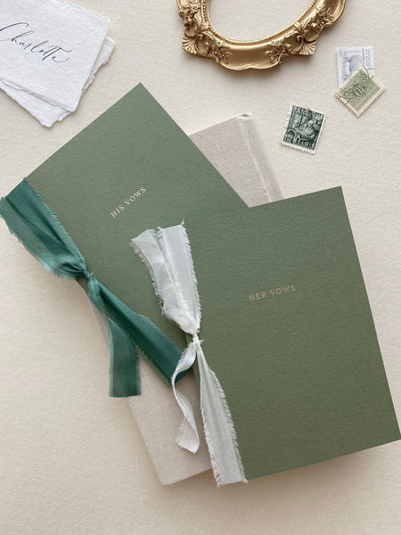 A set of two His and Her gold foil olive green card stock vow books in typeface font with one tied in forest green colored silk ribbon and the other in antique white colored silk ribbon
