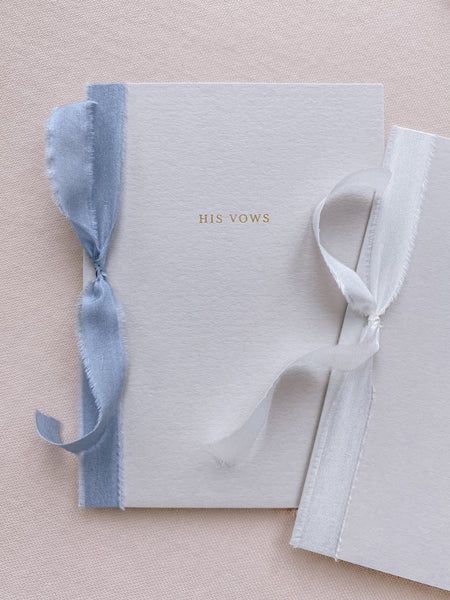 A set of two His and Her gold foil gray card stock vow books in typeface font with one tied in white colored silk ribbon and the other in blue colored silk ribbon
