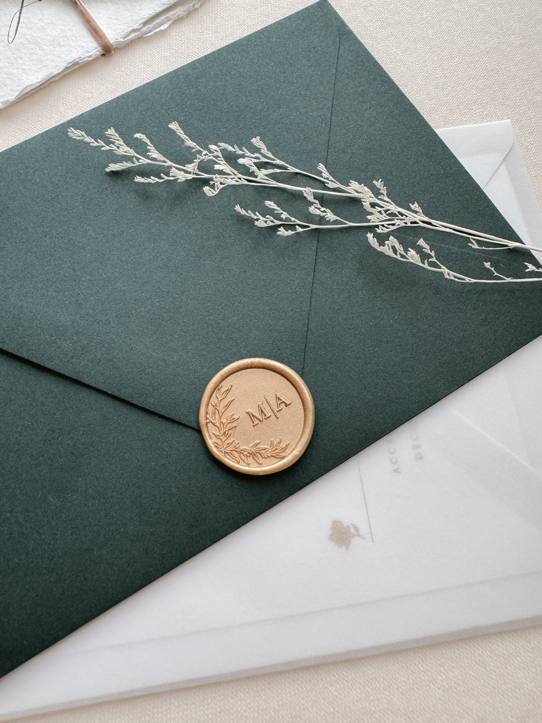 Light gold personalized initials wax seal featuring a custom botanical wreath monogram on a dark green envelope and wedding invitation vellum envelope