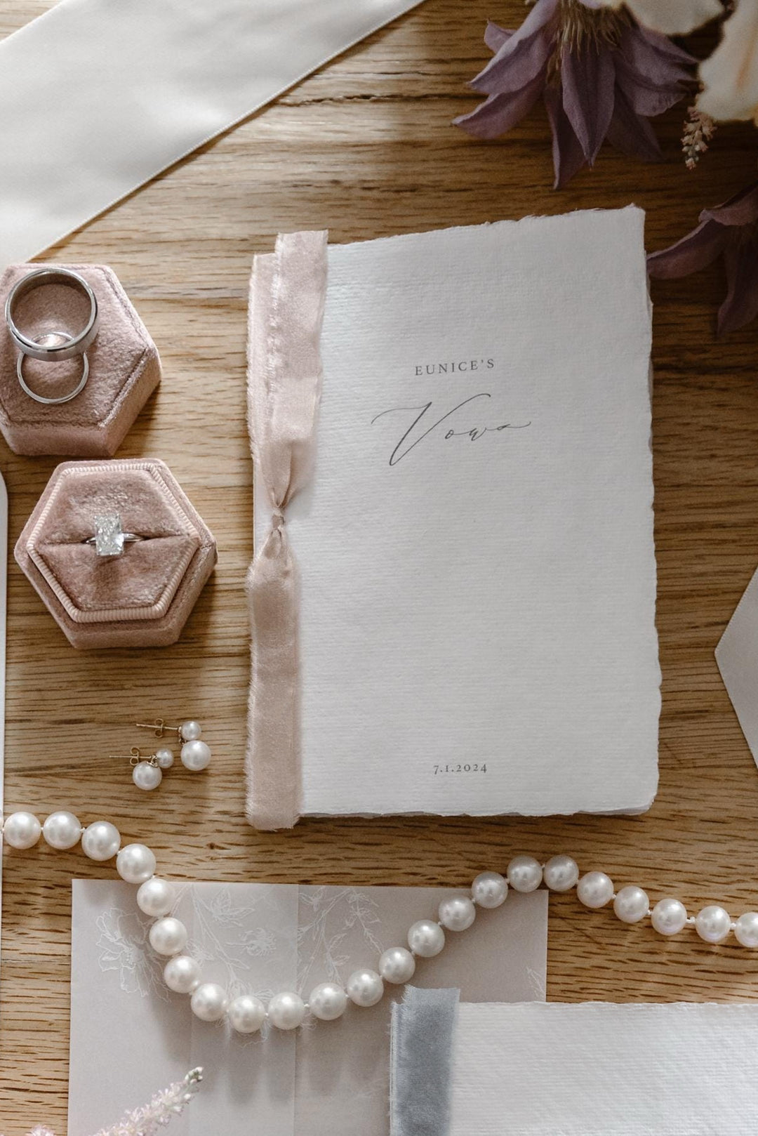 wedding details flat lay showcasing white personalized wedding vow book with pink ribbon 