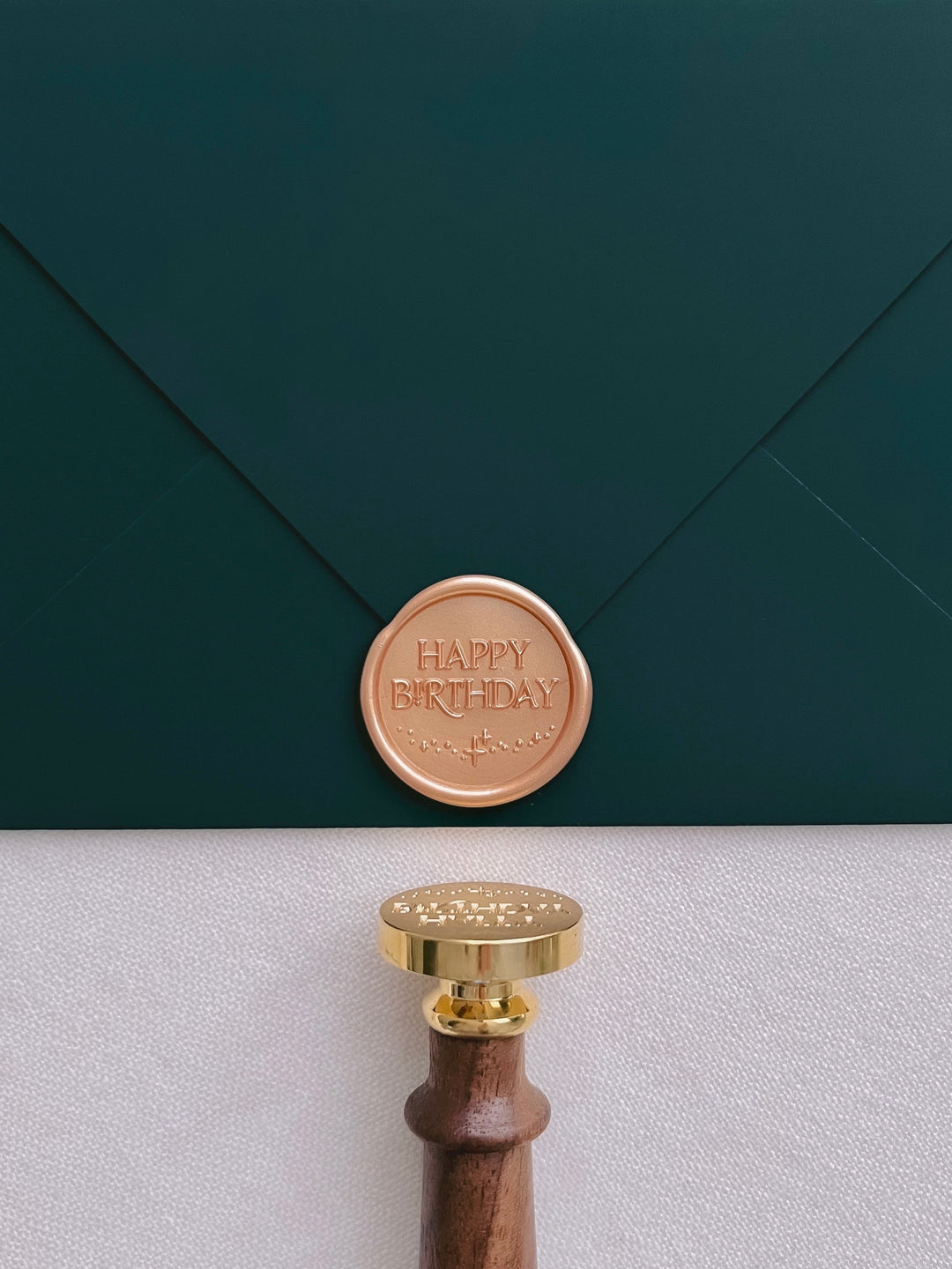 Peachy gold Happy Birthday wax seal on a dark green envelope with a wax stamp head and wooden handle