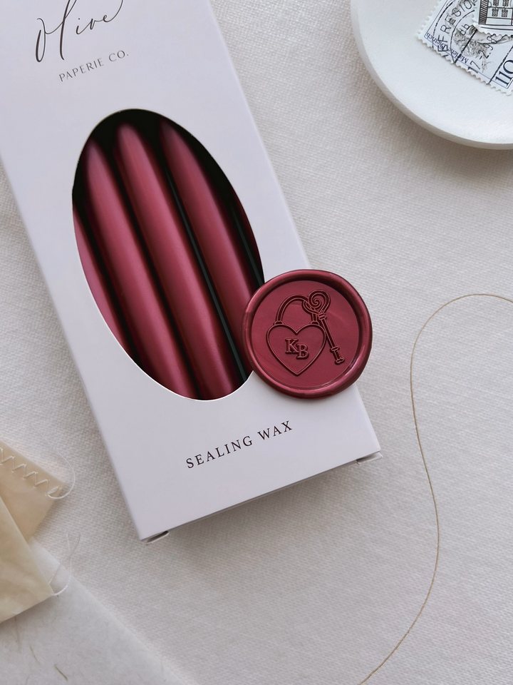 Custom monogram heart shaped lock with key wax seal design featuring personalized initials placed on a box of dark metallic red sealing wax sticks 