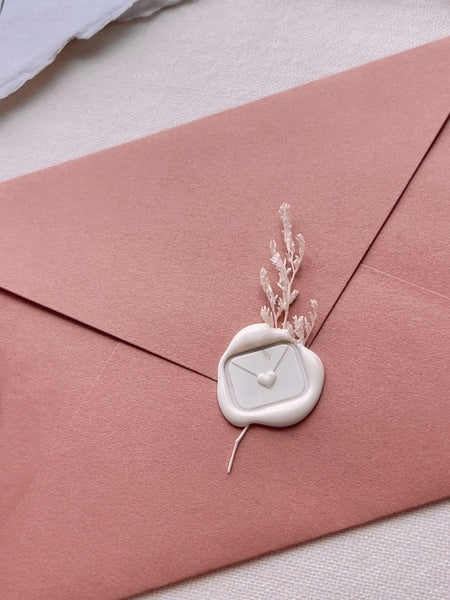 Off-white wax seal featuring an envelope design sealed with a heart, embellished with a white floral branch on a terracotta colored envelope