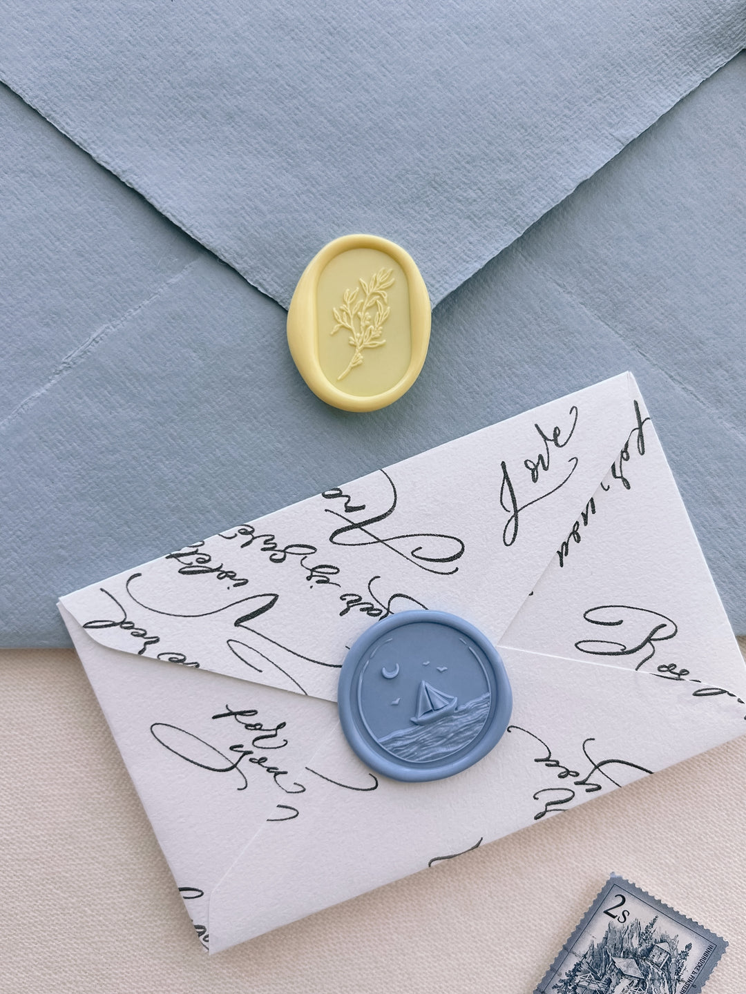 yellow botanical design oval wax seal on a light blue envelope and a blue sailboat wax seal on a white envelope