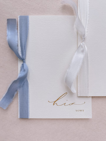 A set of two His and Her gold foil ivory card stock vow books in calligraphy script with one tied in white colored silk ribbon and the other in blue colored silk ribbon