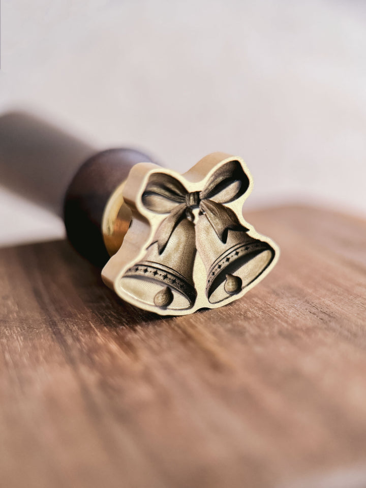 Jingle bells wax seal stamp, showing the die-cut brass stamp head engraved with two bells and a ribbon bow, resting on a wooden surface.