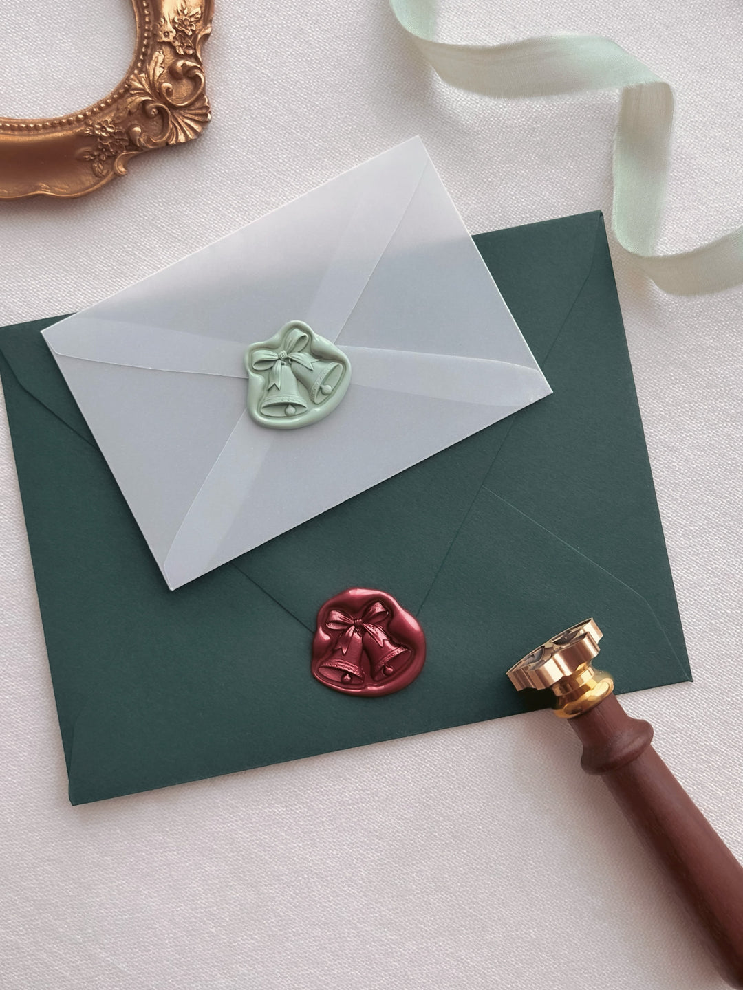 Dark metallic red and light sage green jingle bells 3D die-cut wax seals on a dark green envelope and a mini vellum envelope, styled with a brass wax stamp head with wooden handle, light green silk ribbon and a vintage gold frame 