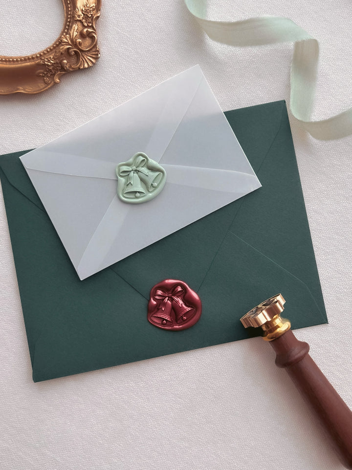 Dark metallic red and light sage green jingle bells 3D die-cut wax seals on a dark green envelope and a mini vellum envelope, styled with a brass wax stamp head with wooden handle, light green silk ribbon and a vintage gold frame 