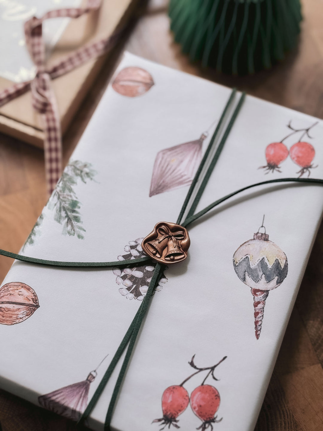 Dark bronze die-cut jingle bells wax seal securing green suede cord on festive illustrated Christmas ornament gift wrap.