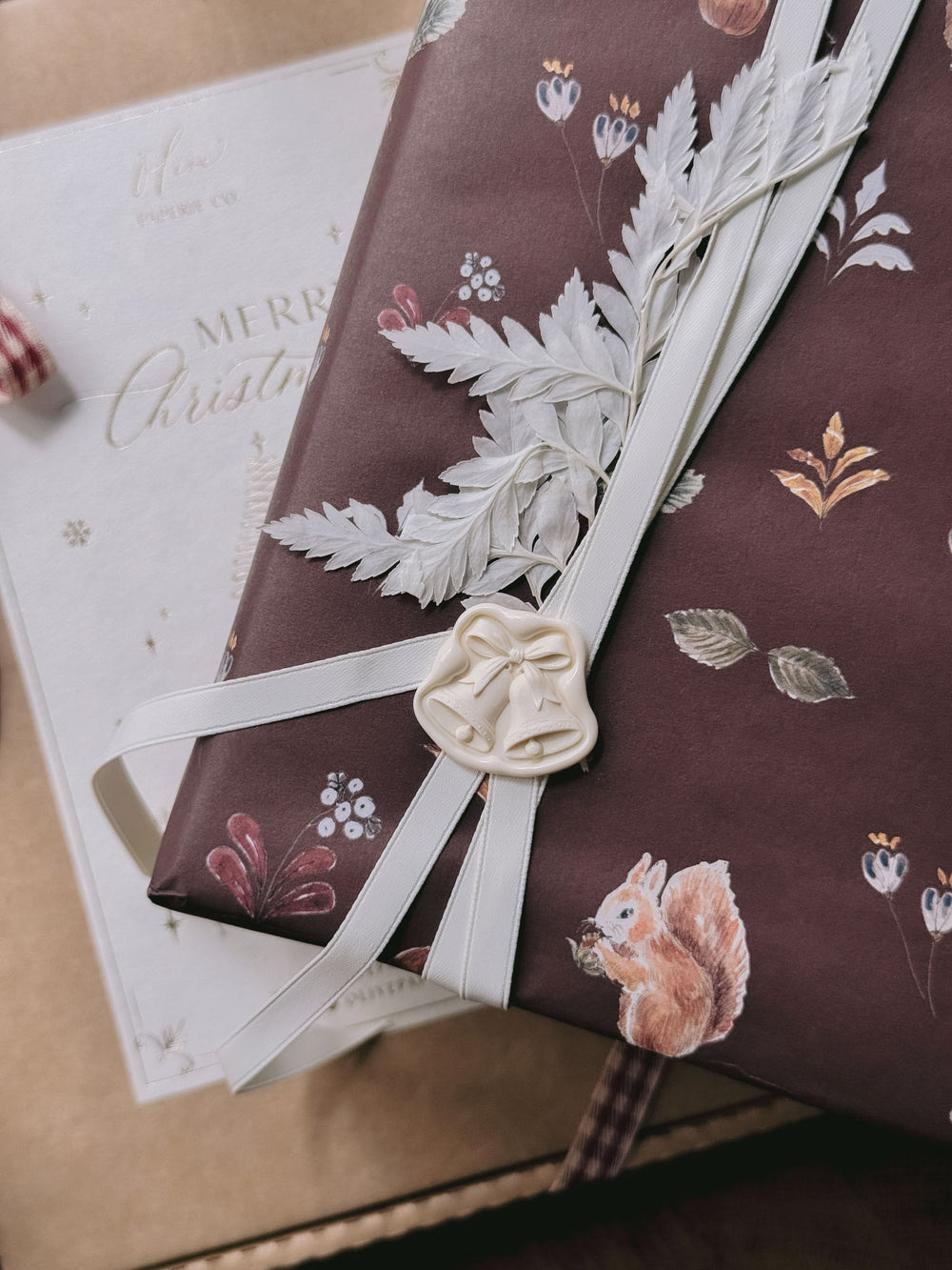 Off-white die-cut jingle bells wax seal on festive burgundy woodland gift wrap, tied with ivory ribbon and dried foliage.