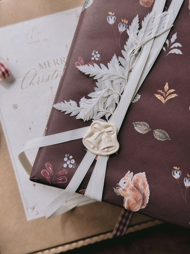 Off-white die-cut jingle bells wax seal on festive burgundy woodland gift wrap, tied with ivory ribbon and dried foliage.