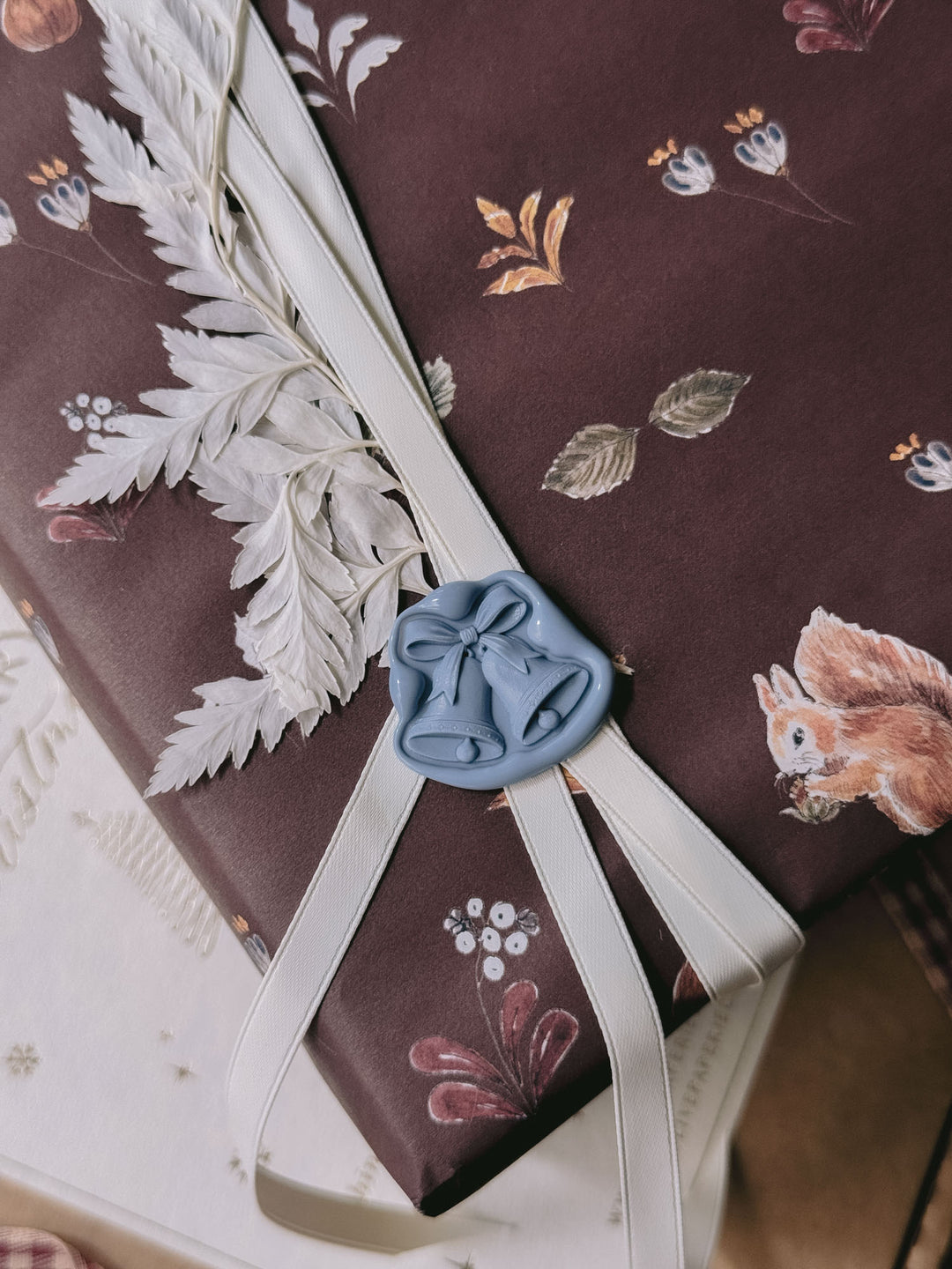 Close-up of a dusty blue die-cut jingle bells wax seal on festive burgundy botanical gift wrap, finished with ivory ribbon and dried white foliage.