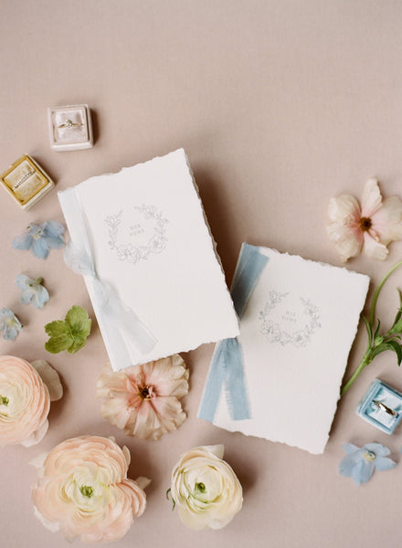 A set of two His and Her letterpress floral wreath vow books with one tied in soft white colored silk ribbon and one tied in pale blue colored silk ribbon styled with flowers and wedding rings