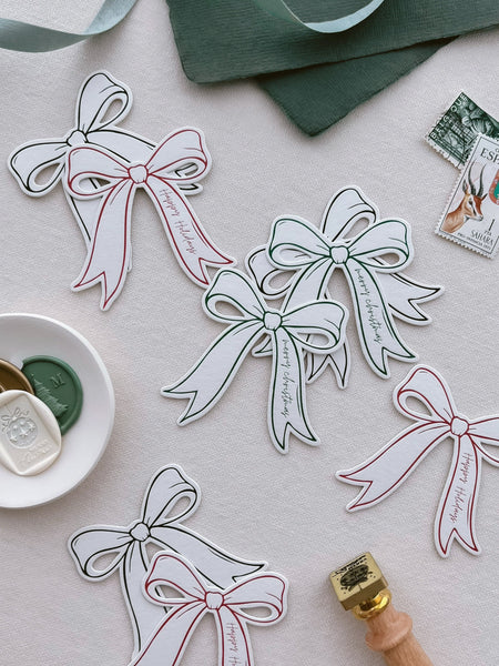 Die-cut bow shaped letterpress gift tags in assorted colors green, red and black. Printed with phrases Merry Christmas and Happy Holidays 