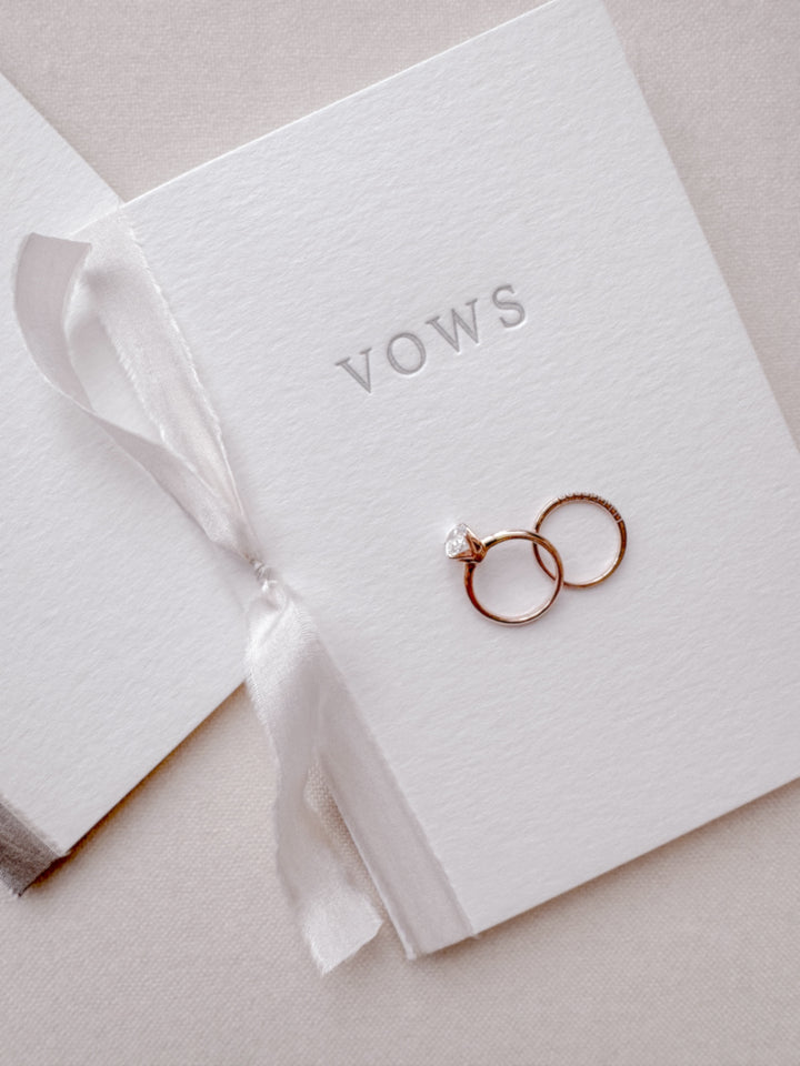 Set of two wedding vow book with VOWS letterpress embossed in gray ink, tied in white silk ribbon and styled with two wedding rings placed on the cover