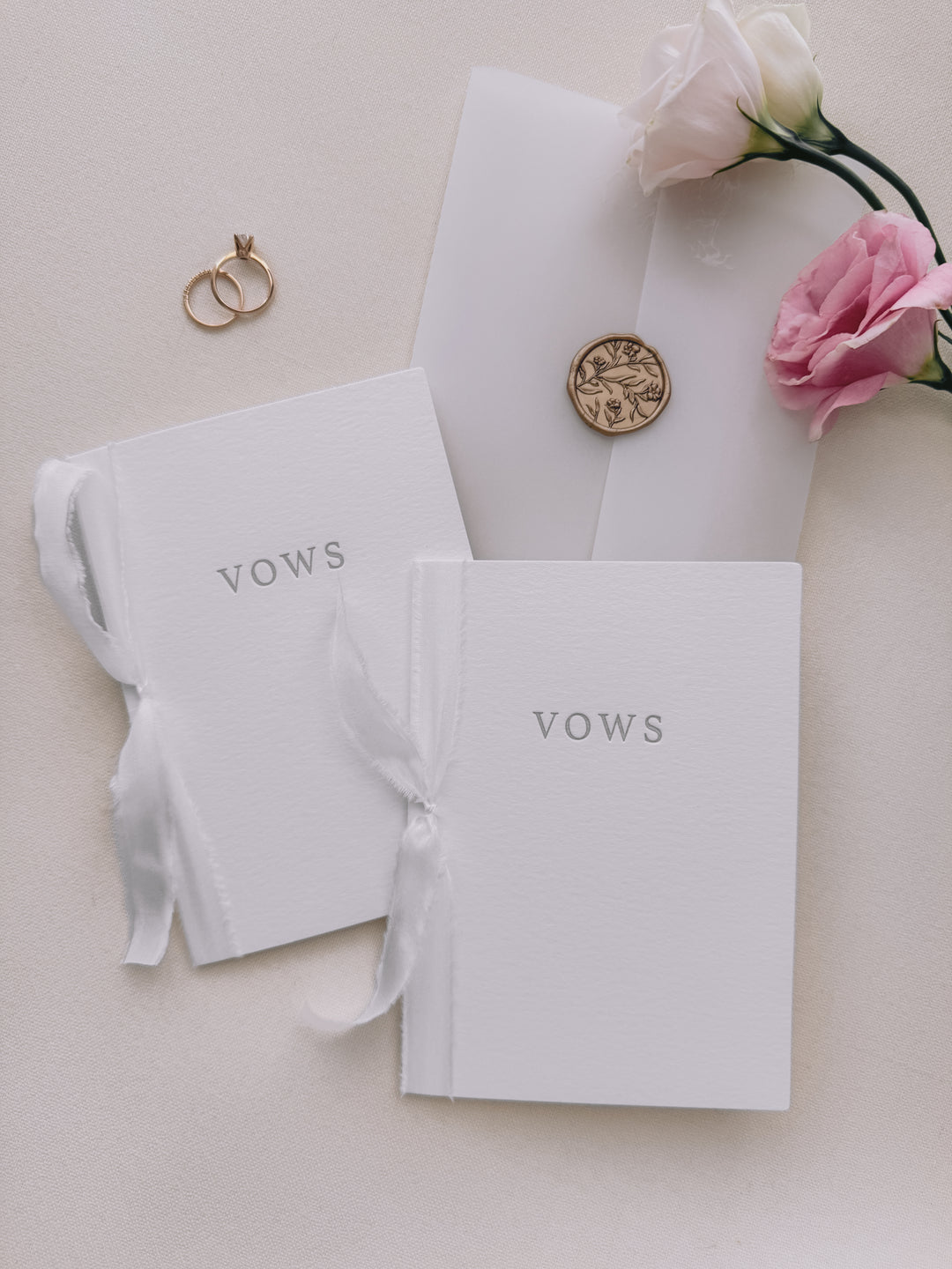 Set of two wedding vow book with VOWS letterpress embossed in gray ink, tied in white silk ribbon and styled with two wedding rings, vellum jacket, a gold floral wax seal and pink flowers