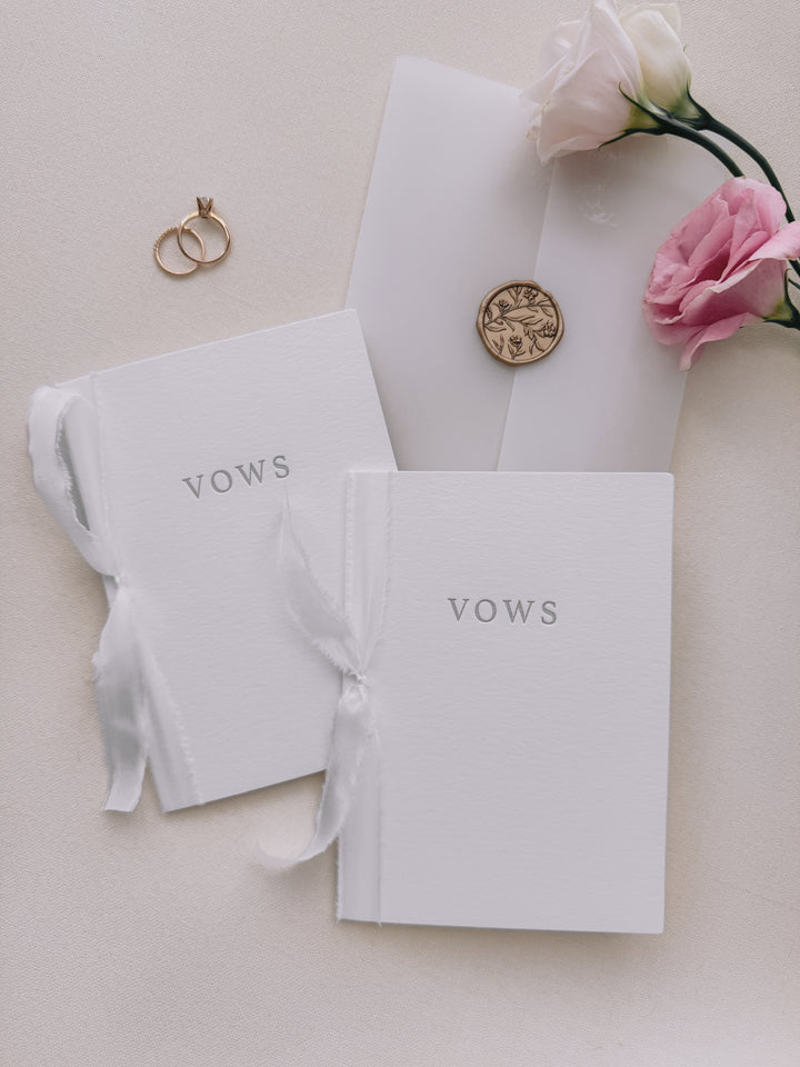 Set of two wedding vow book with VOWS letterpress embossed in gray ink, tied in white silk ribbon and styled with two wedding rings, vellum jacket, a gold floral wax seal and pink flowers
