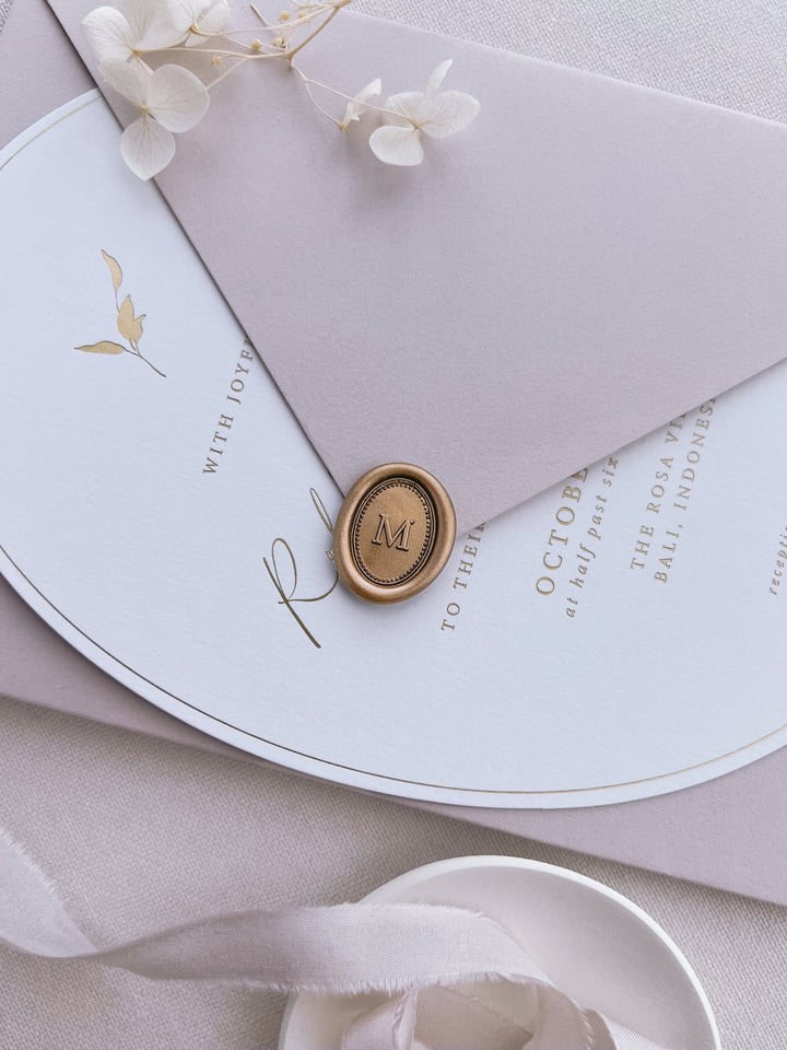 Personalized initial mini oval border gold wax seal featuring a custom monogram on a dusty nude envelope over a gold foil invitation card
