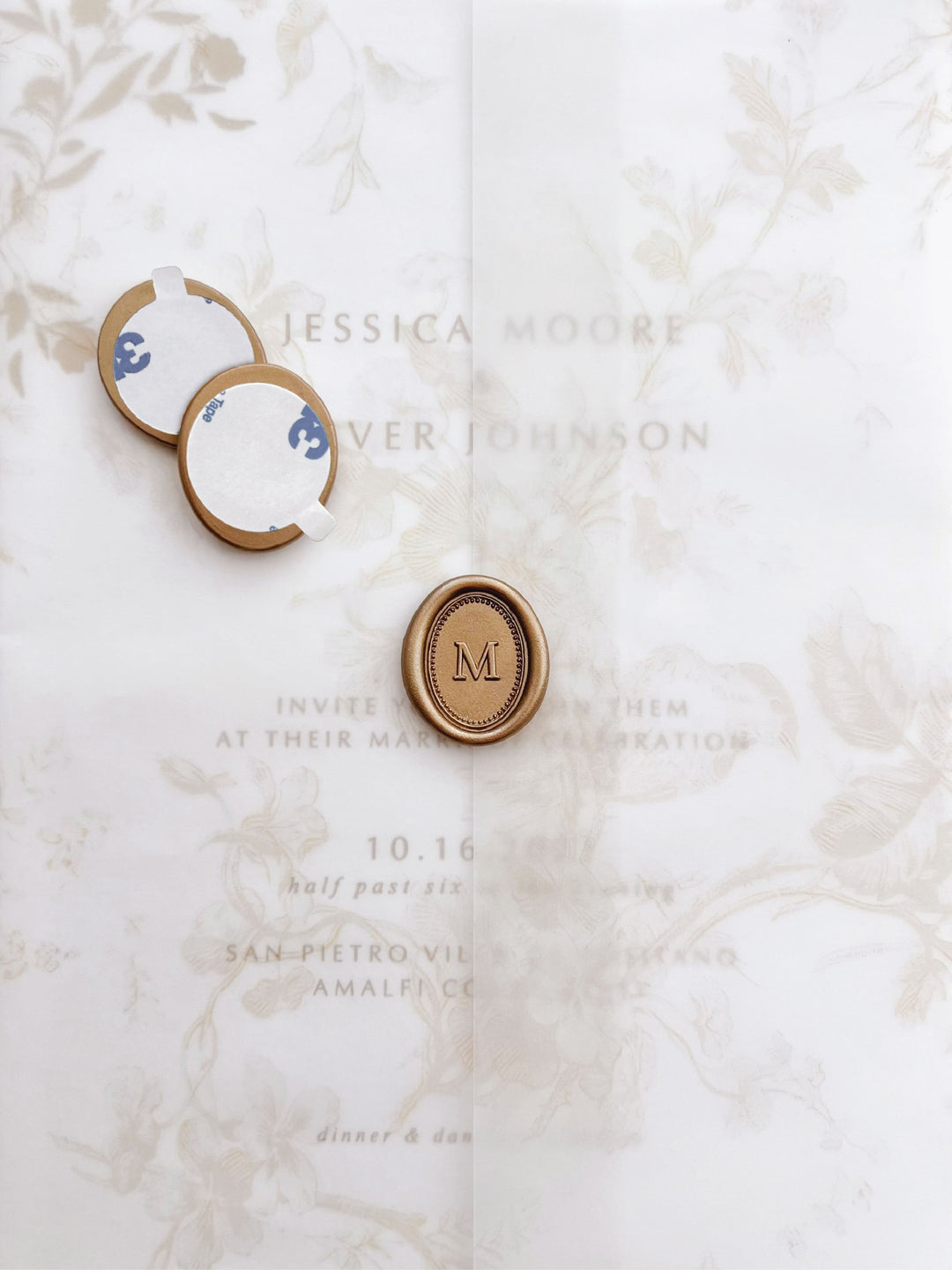 Personalized initial mini oval border gold wax seal stickers with self-adhesive backings on a beige floral vellum jacket over a wedding invitation card 