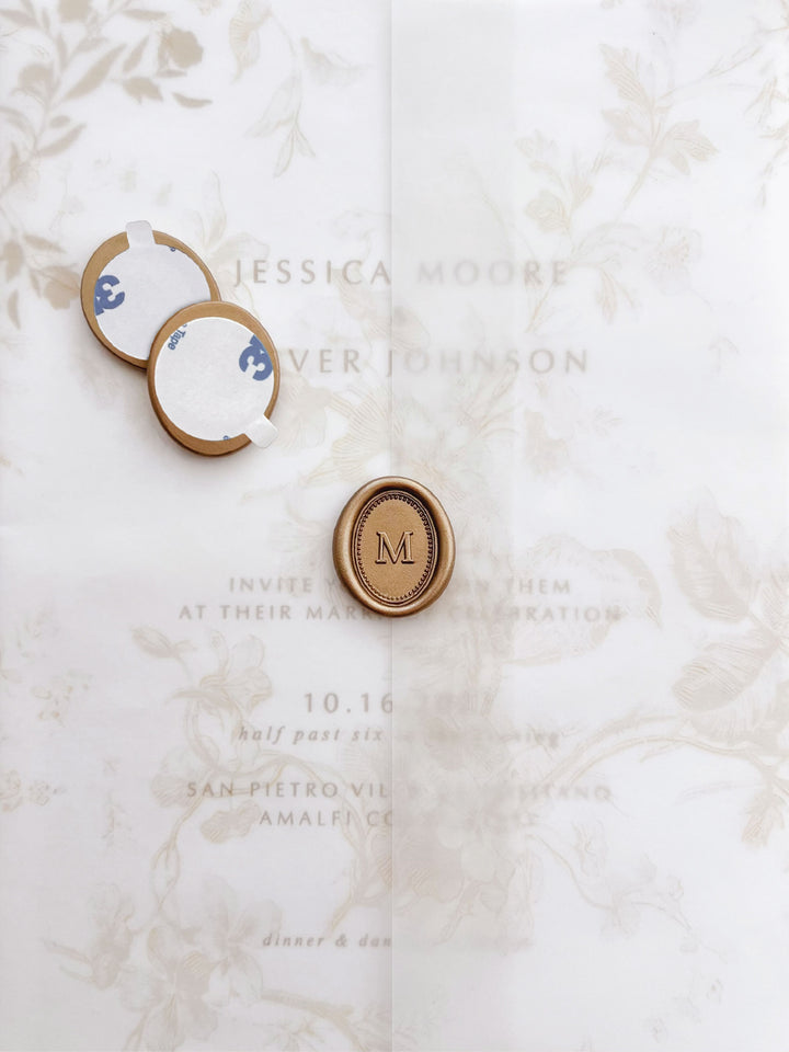 Personalized initial mini oval border gold wax seal stickers with self-adhesive backings on a beige floral vellum jacket over a wedding invitation card 