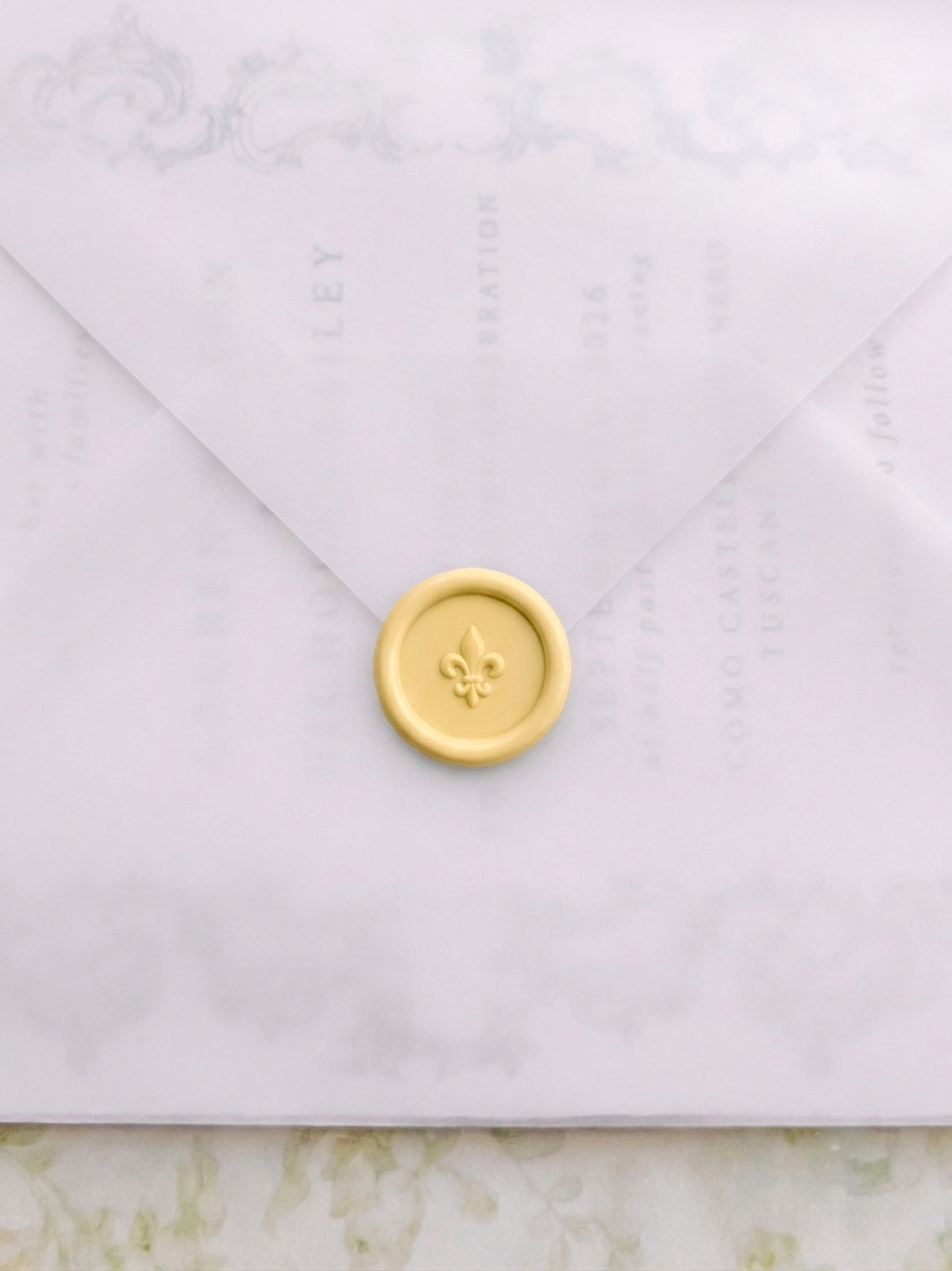 Butter yellow fleur de lis wax seal on a vellum wedding envelope, layered over a botanical wedding invitation suite with delicate scroll detail.