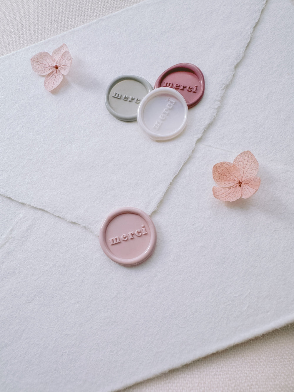 Light pink, antique white, light sage, and rose pink mini merci wax seals on a white deckled edge handmade paper envelope on a light beige background