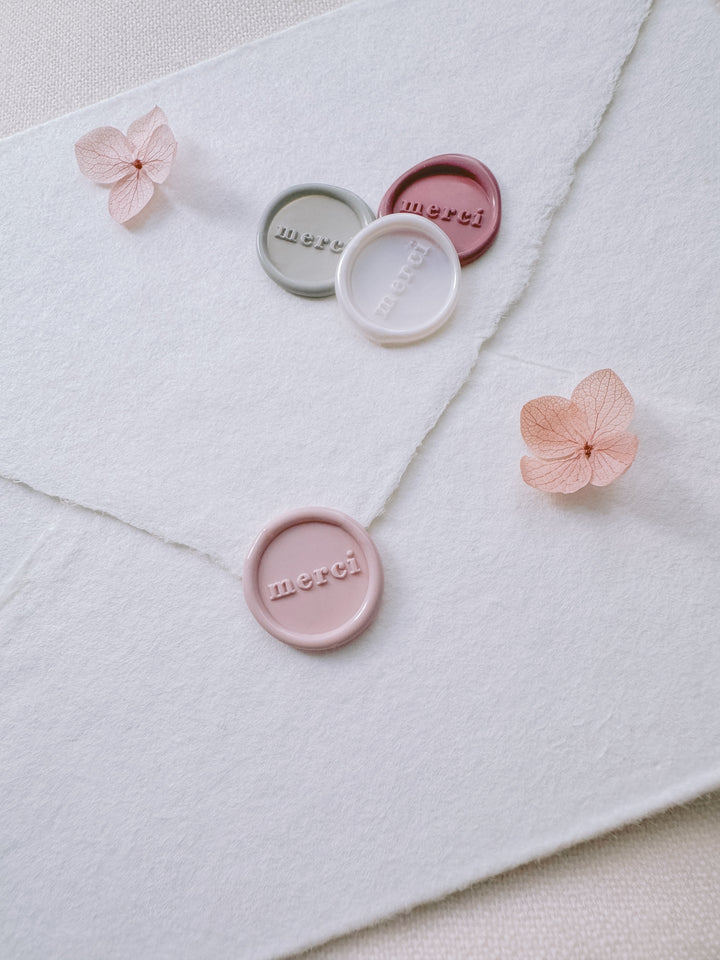 Light pink, antique white, light sage, and rose pink mini merci wax seals on a white deckled edge handmade paper envelope on a light beige background