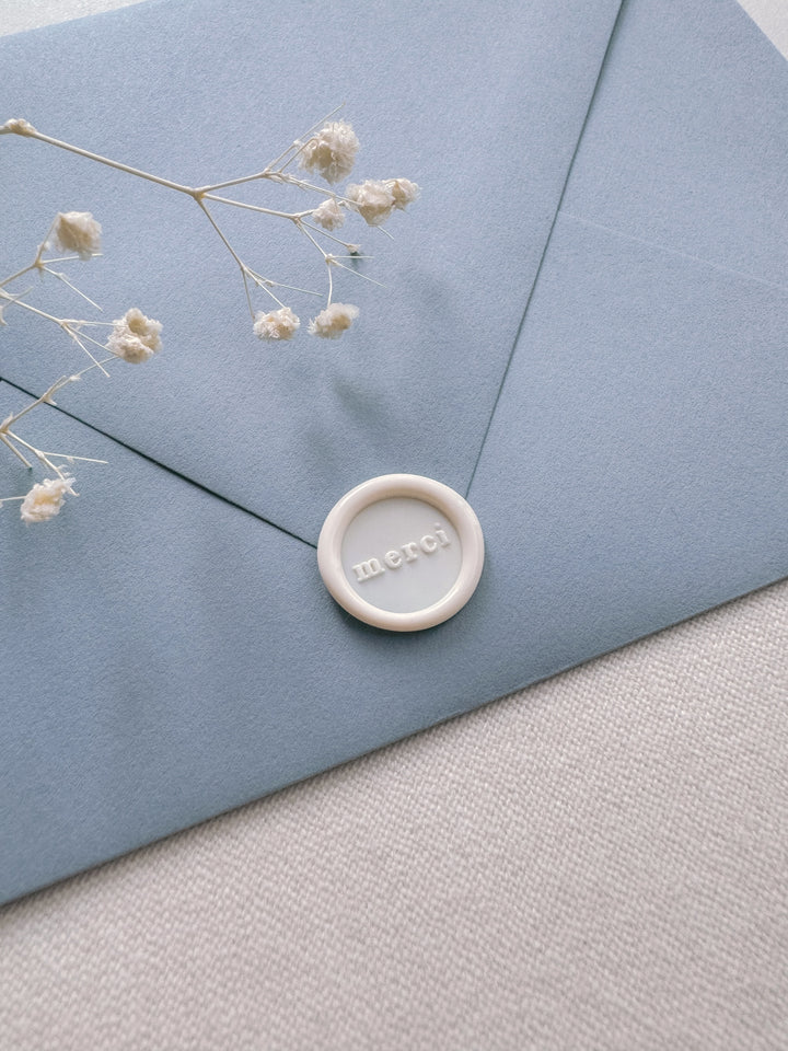 Antique white mini merci wax seal on a dusty blue envelope on a light beige background