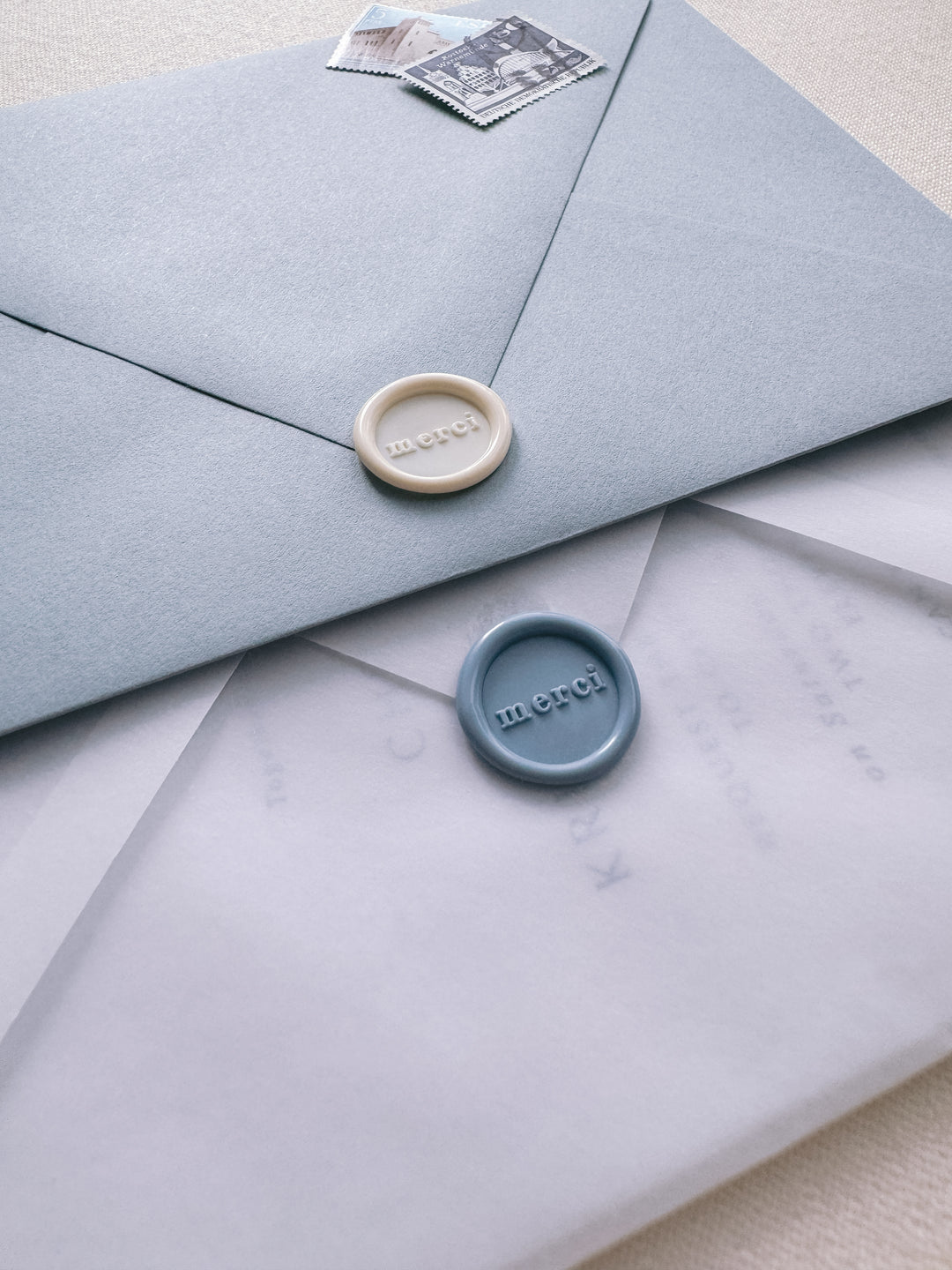Dusty blue mini merci wax seal on a wedding invitation vellum envelope beside an off-white mini merci wax seal on a dusty blue envelope